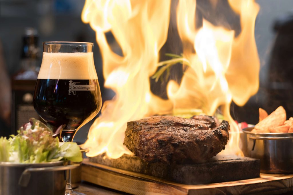 A glass of dark winter beer with frothy top, next to a steak cooking on a grill
