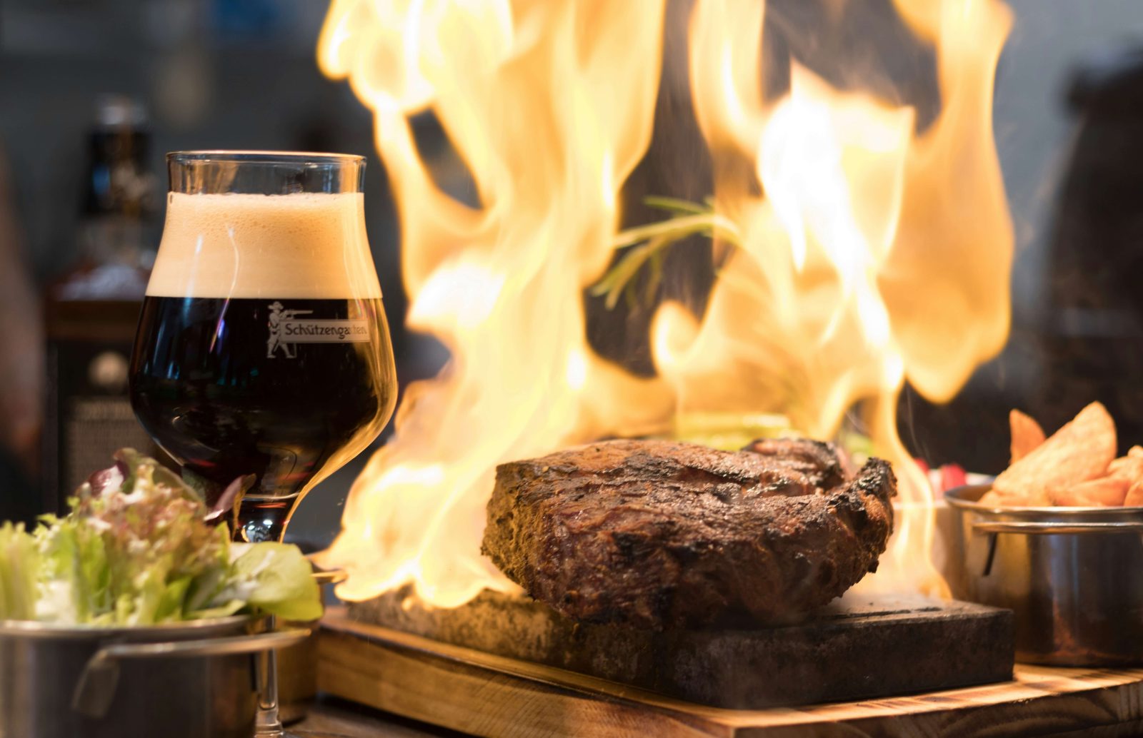 A glass of dark beer with frothy top, next to a steak cooking on a grill