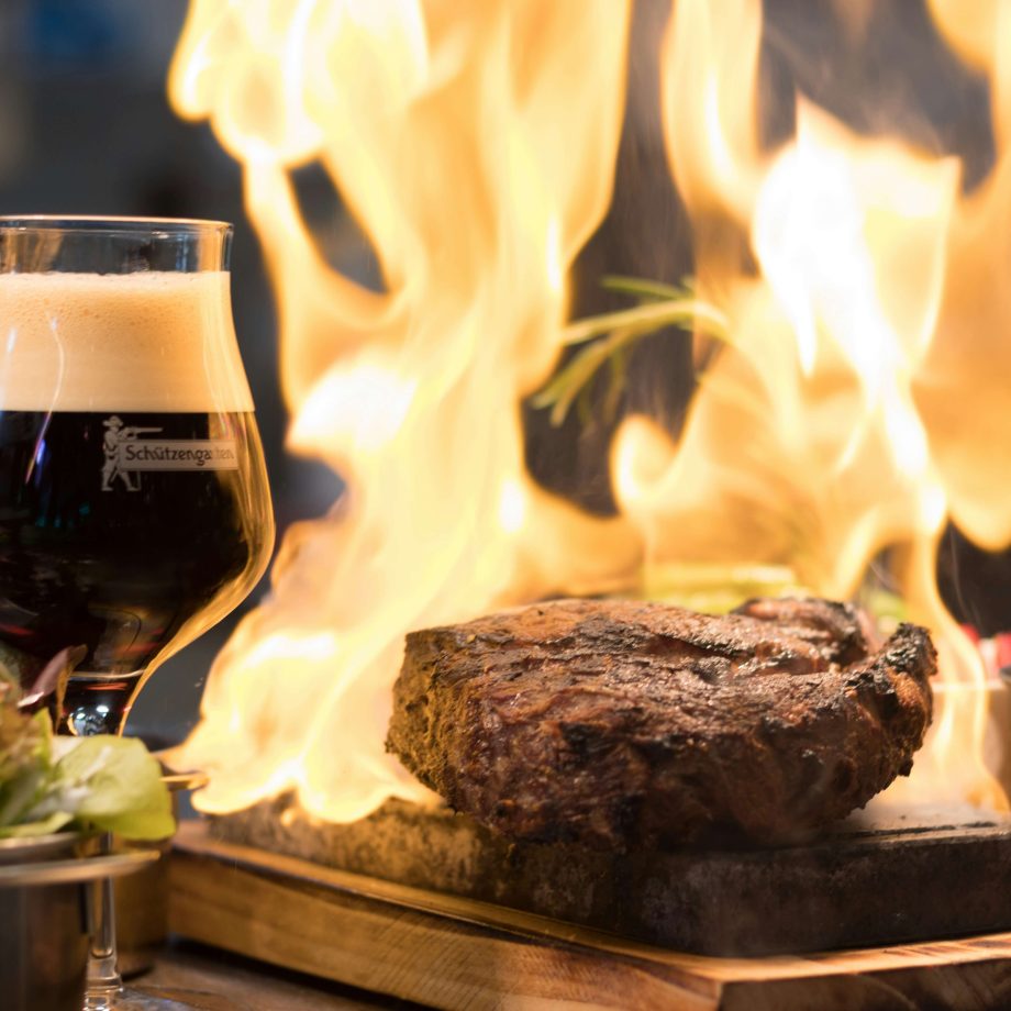 A glass of dark beer with frothy top, next to a steak cooking on a grill